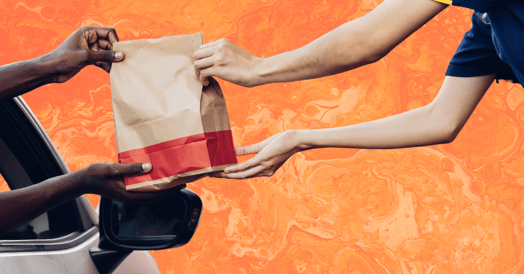 A drive-thru worker hands a paper bag of food to a customer in a car.
